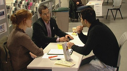 Drei Personen sitzen sich an einem Tisch gegenüber - rechts sitzt ein potenzieller Auszubildender mit schwarzem Pullover, links eine Frau und ein Mann.