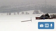 Auf einem einsamen, zugeschneiten Feld liegt ein kleiner, abgeknickter Stommast. Im Hintergrund steht ein kleines rotes Backsteinhaus.