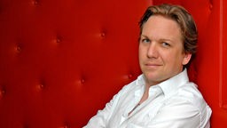 Kabarettist Matthias Brodowy sitzt auf einer roten Couch