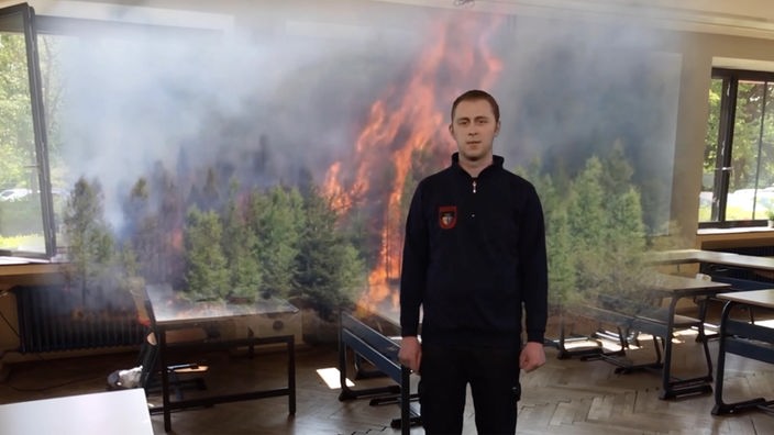 In einen Klassenraum ist virtuell ein Waldbrand projiziert, ebenfalls virtuell steht ein Feuerwehrmann in der Szenerie
