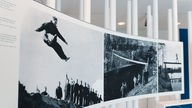 Fotos in einer Ausstellung: Ein junger Mann springt in einen Abgrund, ein junger Mann balanciert über eine Holzstange über einem Abgrund