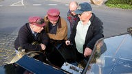 Vier Männer unterhalten sich ernsthaft neben einer geöffneten Motorhaube stehend