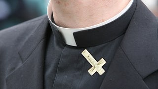 Montage mit Halspartie, weißem Kragen und Schulter eines Priesters, an dessen Revers ein nach unten weisendes kleines Kruzifix befestigt ist