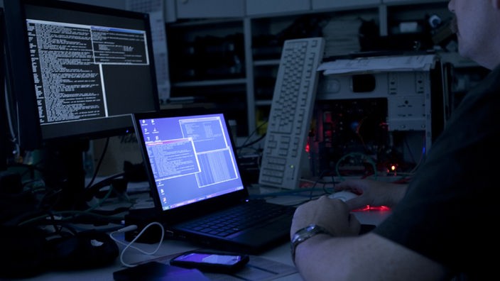 Ein Mann sitzt in einem abgedunkelten Raum vor verschiedenen Computern und Monitoren