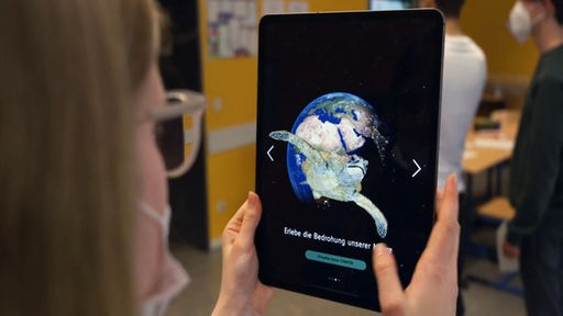 Blick über die Schulter einer Schülerin, die ein Tablet in den Händen hält. Auf dem Tablet sind eine Weltkugel und eine Schildkröte zu sehen.
