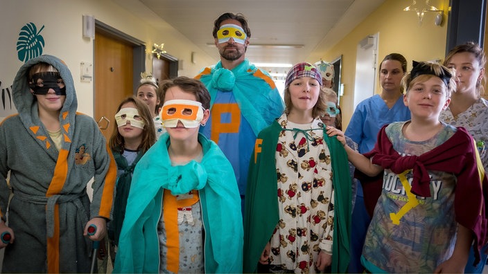 Schwester Carola (Anna König, re.) freut es: Die Kinder auf der Kinderstation ziehen mit Pohlmann (Benjamin Sadler, Mitte) als Superhelden umher. Mit dabei Benny (Caspar Gierden, li.), Emma (Hannah Franke), Paul (Oskar Netzel, 3. v. li.).