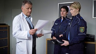 Dr. Roland Heilmann (Thomas Rühmann, li.) wird während einer Operation von der Polizei (Anke Stoppa, re., Komparse, 2.v.re.) zu einem dringenden Gespräch in sein Büro gerufen.