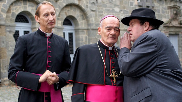 Pfarrer Braun (Ottfried Fischer, re.) verrät Monsignore Mühlich (Gilbert von Sohlern, li.) und Bischof Hemmelrath (Hans-Michael Rehberg) ein Geheimnis.