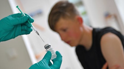 Eine Impfung wird vorbereitet: Im Vordergrund die Nadel, im Hintergrund ein Jugendlicher mit hochgerolltem Ärmel