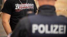 Die unscharfe Rückenansicht einer Polizeianform vor einer Person, die ein Shirt mit einer Aufschrift in altdeutscher Schrift trägt 