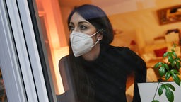 Dunkelhaarige Frau mit FFP2-Maske schaut aus dem Fenster