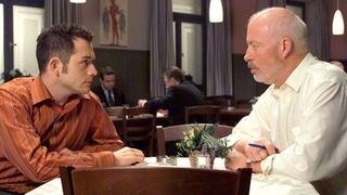 Erich (Bill Mockridge, rechts) erfährt von Alex (Joris Gratwohl), dass Helga die Arbeitsaufteilung im Reisebüro offensichtlich ohne ihn plant.