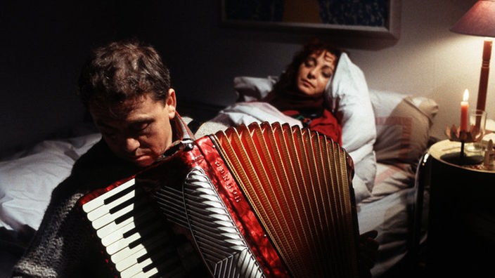 Olaf (Franz Rampelmann) versucht seine ans Bett gefesselte Frau (Manon Straché) musikalisch aufzuheitern.