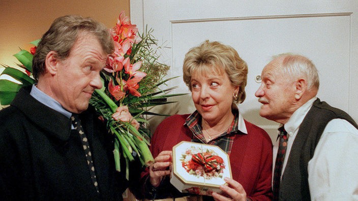 Onkel Franz (Martin Rickelt, re) bekommt mit, wie sich Lösch (Wichart von Roell) mit Blumenstrauß und Naschwerk bei Helga (Marie-Luise Marjan) ins Zeug legt.