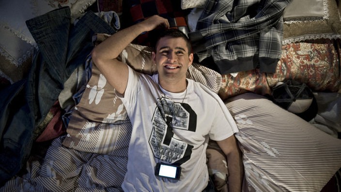Orkan (Hüseyin Ekici) liegt freudestrahlend mit seinem Handy auf dem Bett. Offensichtlich hat er eine schöne Nachricht erhalten.