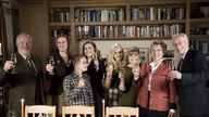 Gruppenfoto von Erich, Anna, Sarah, Martin, Lea, Sophie, Helga und Hans im Wohnzimmer. Sie prosten dem Zuschauer  mit Sektgläsern zu.