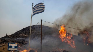 Flammen lodern auf einem Hügel und es ist eine Griechenland-Flagge zu sehen.