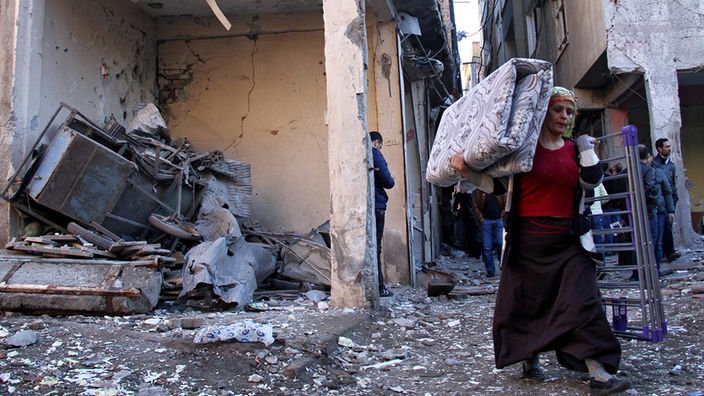 Junge Frau trägt eine Matratze und einen Wäscheständer durch eine zerstörte Straße