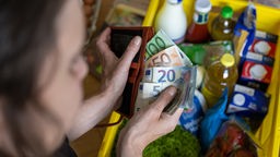 Eine Frau hält Geldscheine in einer Geldbörse über einer Einkaufskiste mit Lebensmitteln.