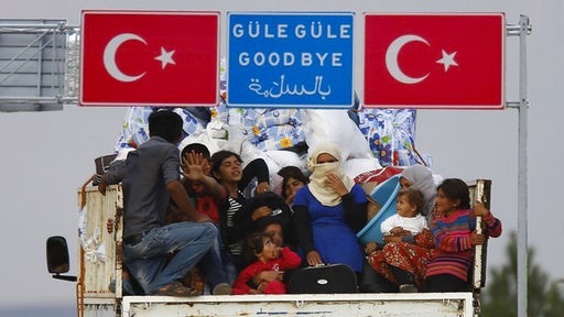 Offener Lastwagen mit Flüchtlingen unter einem türkischen Autobahnschild mit der Aufschrift Güle Güle, Goodbye