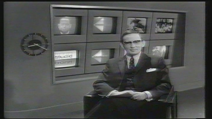 Ein Moderator sitzt auf einen Sessel vor einer Wand mit 8 Fernsehern