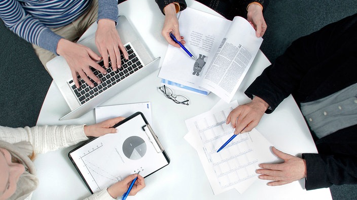 Ein Redaktionsteam (bestehend aus vier Personen) sitzt an einem runden Tisch. Es sind nur die Hände zu sehen. Auf dem Tisch befinden sich ein Laptop und mehrere Papierunterlagen für die Redaktionsbesprechung. Aufnahme von oben.