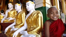 Ein Kind in einer roten Mönchsrobe steht lächelnd vor einigen Buddhastatuen