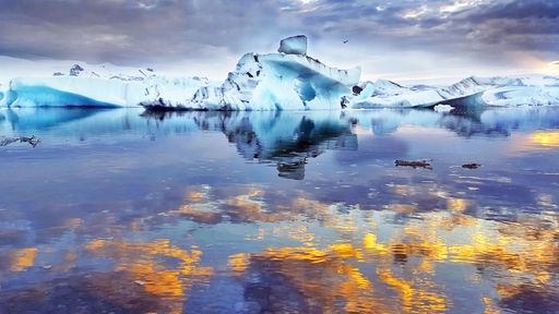 Ein See mit einem Eisberg und im Wasser spiegelt sich die Mitternachtssonne in Goldtönen.