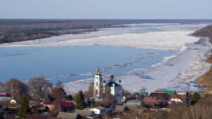 Eine Luftaufnahme der sehr breiten Wolga mit Blick auf Fluss und einem Dorf mit Kirche im Winter.
