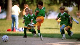 Kinder spielen Fußball in einem Park. Ein Junge im grünen Trikot schießt den Ball.