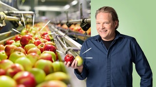 Björn Freitag hält zwei Äpfel in seiner Hand und lächelt in die Kamera. Im Hintergrund ist ein Fließband mit verschiedenen Äpfeln zu sehen.