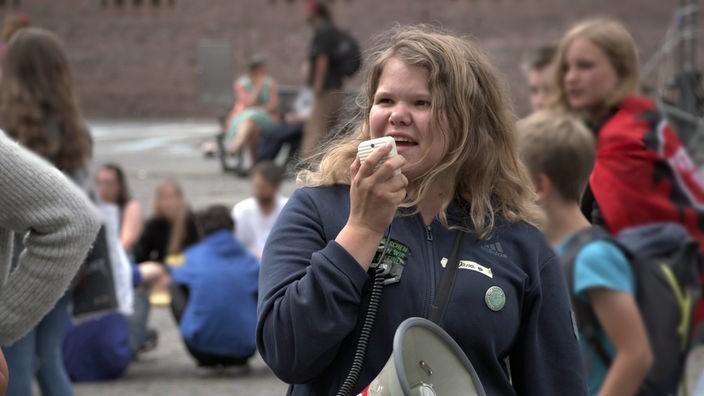 Eine Jugendliche spricht auf der Straße in ein Megafon. Im Hintergrund sind weitere Jugendliche zu sehen.