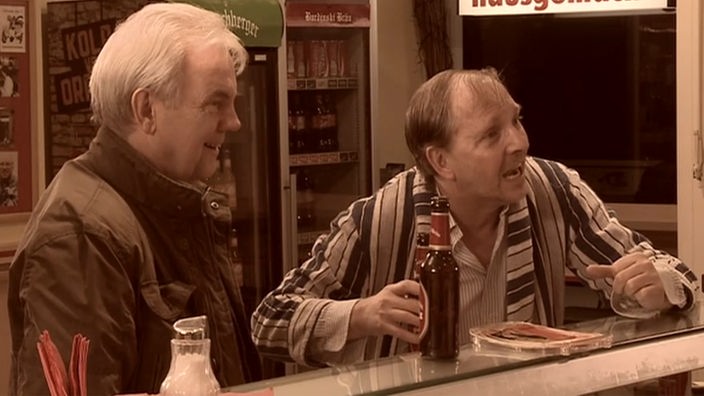 Stammgast Knut Hartmann und Dittsche stehen am Tresen in der Eppendorfer Grillstation, beide lachen und halten ein Burdinski Bräu-Bier in der Hand