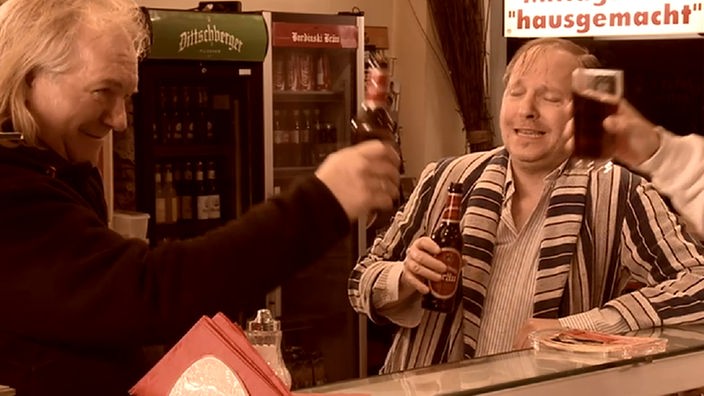 Jens Lindschau hält eine Bierflasche, Ingo ein Colagals im Hintergrund Dittsche mit Bier und geschlossenen Augen.