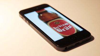 Ein Handy mit einer Bierflasche auf dem Display. Aus dem Handy fliegt ein "Plopp"-Schriftzug 