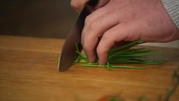 Das Bild zeigt wie Schnittlauch mit einem Messer auf einem Brett geschnitten wird.