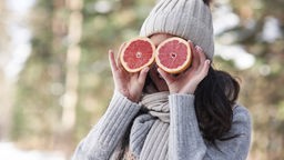 Das Bild zeigt eine Frau mit Schal und Mütze, die sich zwei Grapefruit-Hälften an die Augen hält.