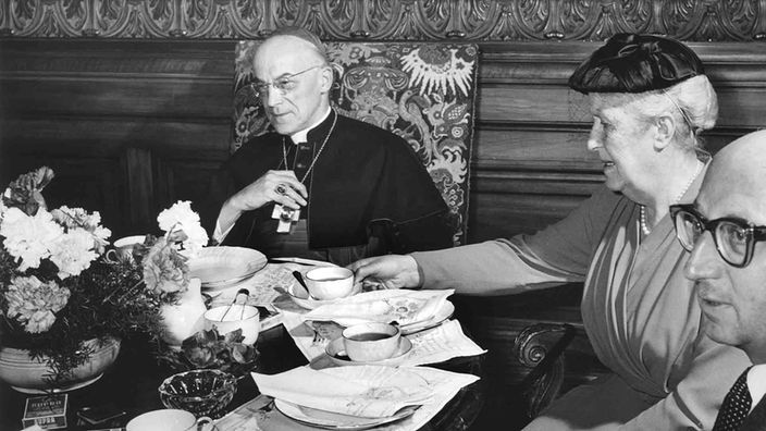 sw: Eine Frau sitzt zwischen einem Kardinal und einem anderen Mann am gedeckten Kaffee-Tisch.