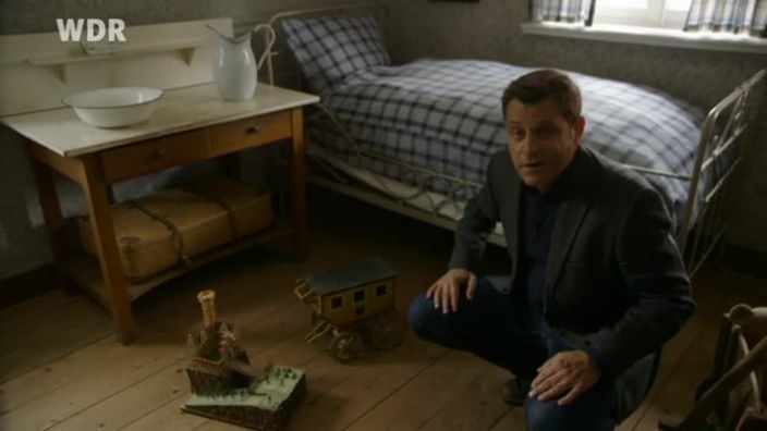 Mann hockt auf dem Boden eines alten Zimmers; vor ihm liegt altes Kinderspielzeug
