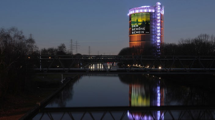 Blick auf das Gasometer Oberhausen bei Nacht mit Plakat zur Ausstellung "Der schöne Schein"