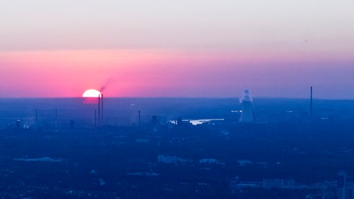 Die Sonne geht rot glühend am Horizont unter, im Vordergrund Industrieanlagen.