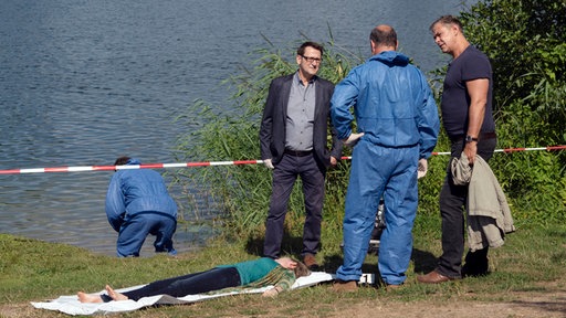 Am Ufer der Trave wurde eine Frauenleiche (Lisa Karlström, liegend) aufgefunden. Lars Englen (Ingo Naujoks, l.), Dr. Strahl (Christoph Tomanek, M.) und Finn Kiesewetter (Sven Martinek, r.) besprechen erste Eindrücke am Tatort.