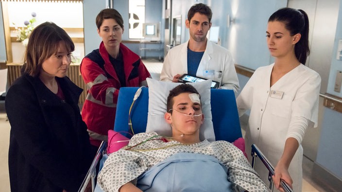 Ein junger Mann liegt auf einem Krankenhausbett, um ihn herum stehen eine Ärztin, ein Arzt und zwei Frauen