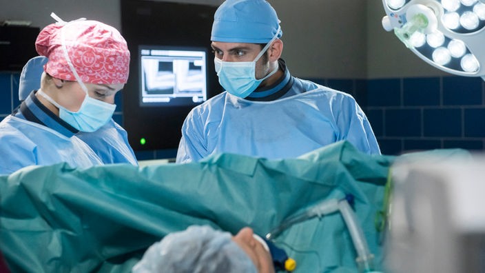Zwei Menschen mit Kopfhaube und Mundschutz stehen vor einem Operationstisch.