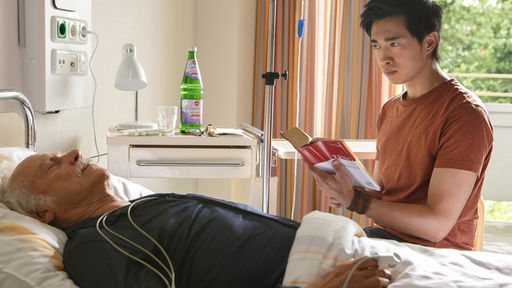 Nach seinem Herzinfarkt liegt Edwin (Tilo Prückner, l.) im Krankenhaus. Neben ihm sitzt Hui Ko (Aaron Le, r.).