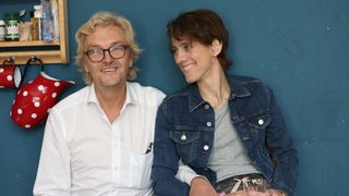Ein lächelndes Paar sitzt nebeneinander vor einer blauen Wand. Der Mann trägt ein weißes Hemd und eine Brille, die Frau eine Jeansjacke. Im Hintergrund ist ein Regal mit Tassen und anderen Gegenständen zu sehen.