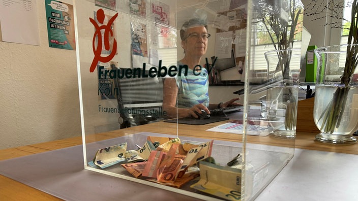 Geldspendebox aus Plexiglas im Vordergrund, Frau am Schreibtisch dahinter