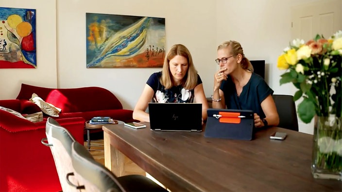 Zwei Frauen sitzen im Wohnzimmer an einem Holztisch und schauen auf ihre Computerbildschirme.