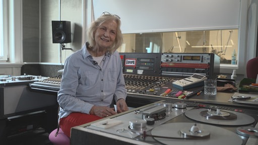 Eine lächelnde Frau mit Brille auf dem Kopf sitzt im Tonstudio vor einem analogen Tonbandgerät.