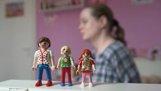 Drei Playmobile Figuren stehen auf Tisch. Frau im Hintergrund unscharf zu erkennen